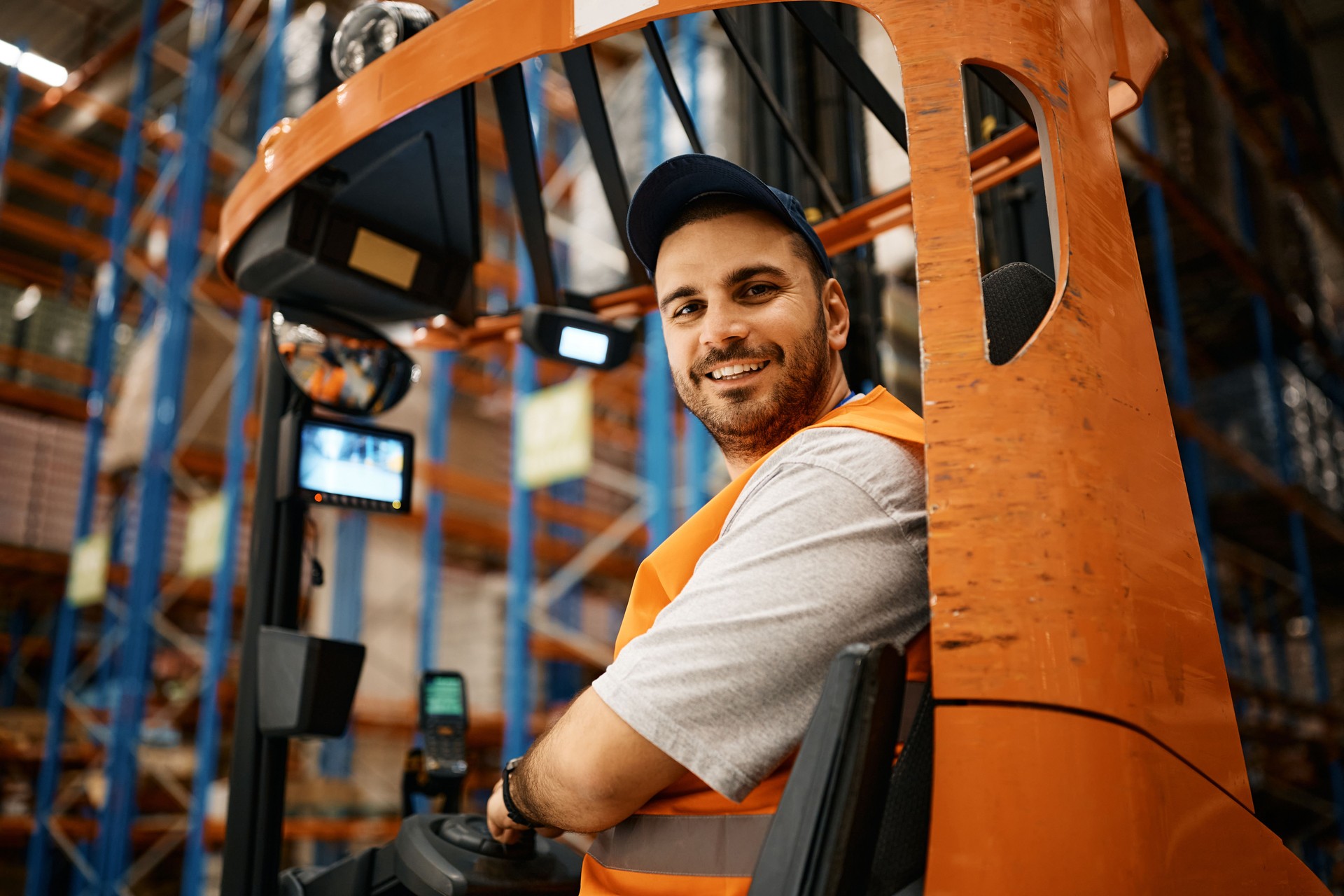 Happy male worker driving forklift at distribution warehouse and looking at camera.